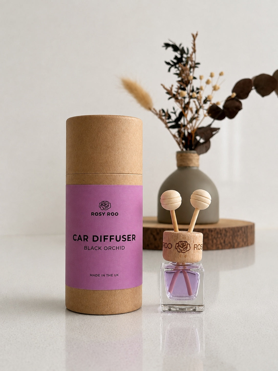 Rosy Roo Black Orchid car diffuser in pink-labeled packaging beside a glass fragrance bottle with wooden reeds.