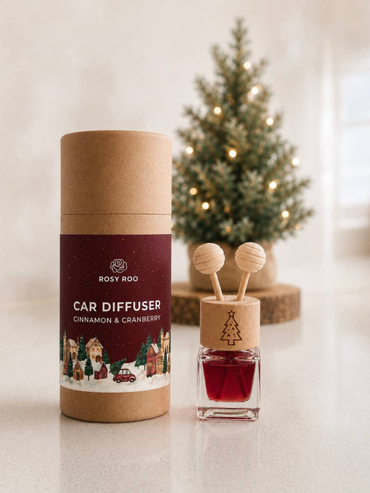 Rosy Roo Car Diffuser in Cinnamon & Cranberry scent, shown with its brown cardboard packaging cylinder and the open diffuser bottle with wooden top and red liquid, set on a light countertop with a blurred Christmas tree in the background.