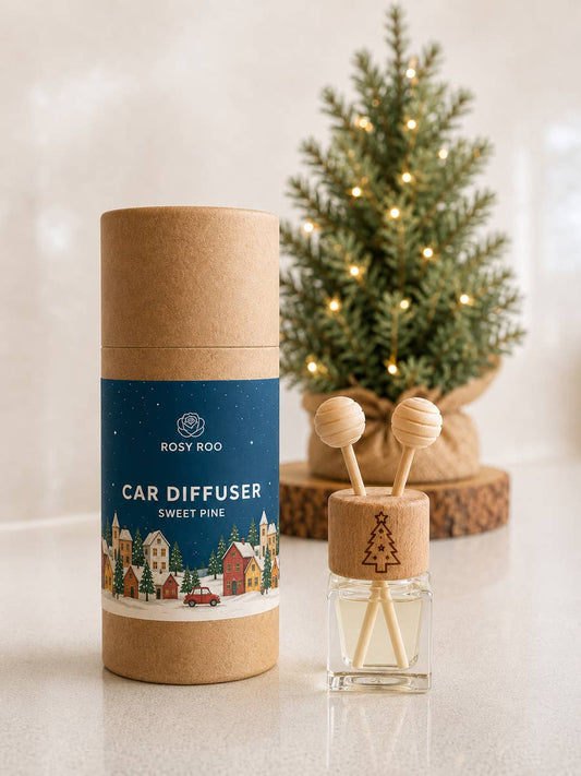 Rosy Roo Sweet Pine Car Diffuser shown with its kraft paper packaging cylinder in the foreground, and the clear glass diffuser bottle with wooden stick tops beside it. A small lit Christmas tree sits blurred in the background on a light surface.