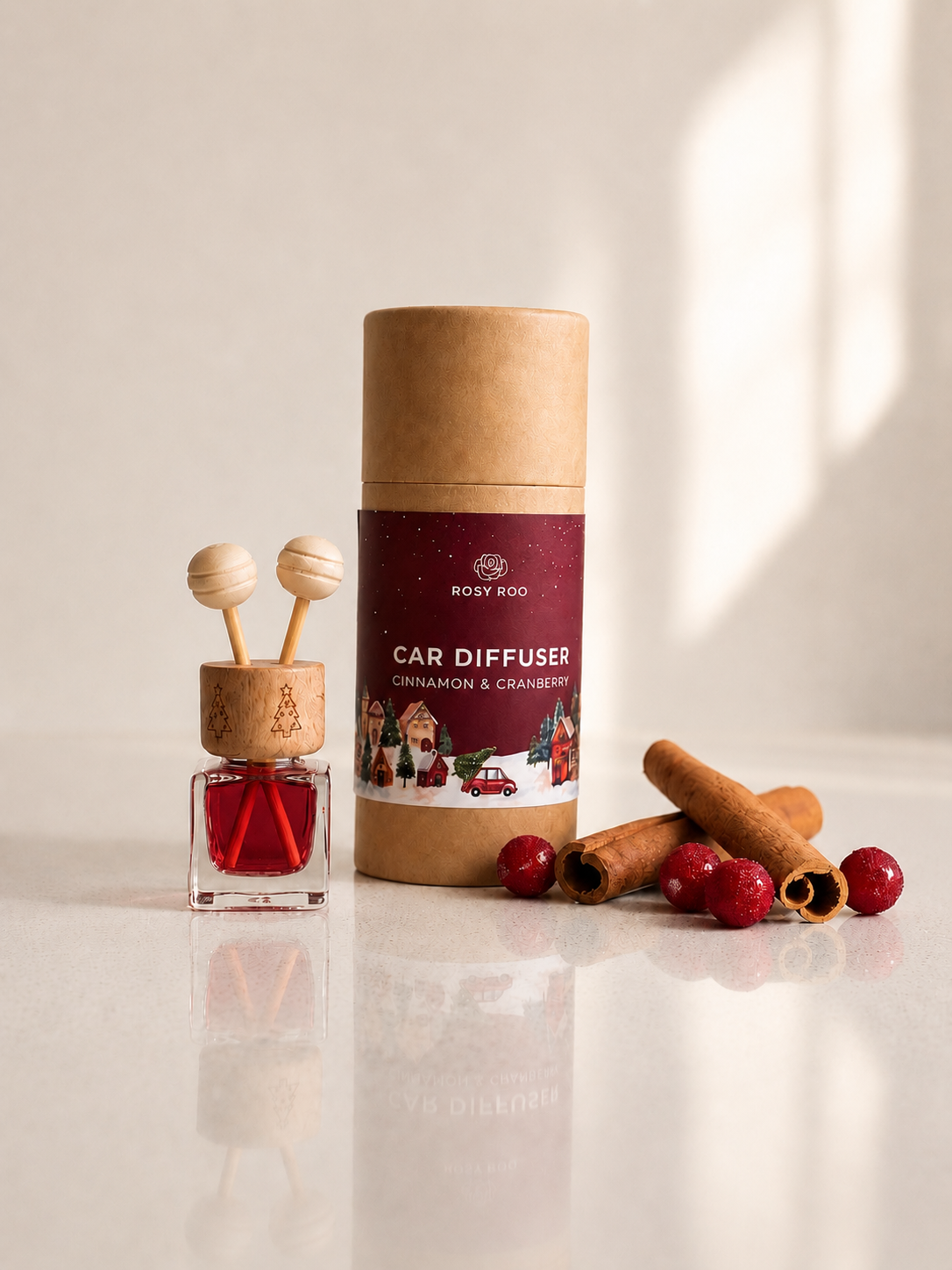 Body 400 Car Diffuser in Cinnamon & Cranberry scent, shown with its kraft paper packaging cylinder, a glass bottle with red liquid and wooden diffuser sticks, and decorative cinnamon sticks and cranberries on a light reflective surface.