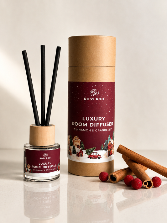 Rosy Roo Luxury Room Diffuser in Cinnamon & Cranberry scent, shown with its brown cardboard cylinder packaging, a glass bottle with black reeds, and decorative cinnamon sticks and cranberries on a light beige background.