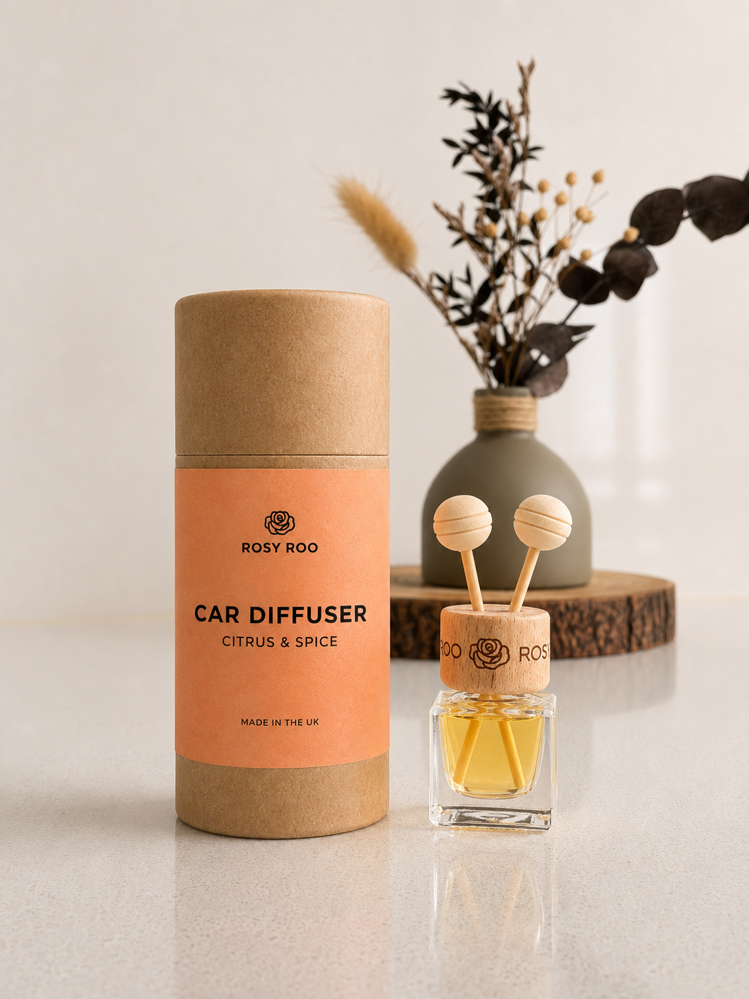 Rosy Roo Citrus & Spice car diffuser in a coral-and-tan package beside its glass oil bottle and decorative reeds.