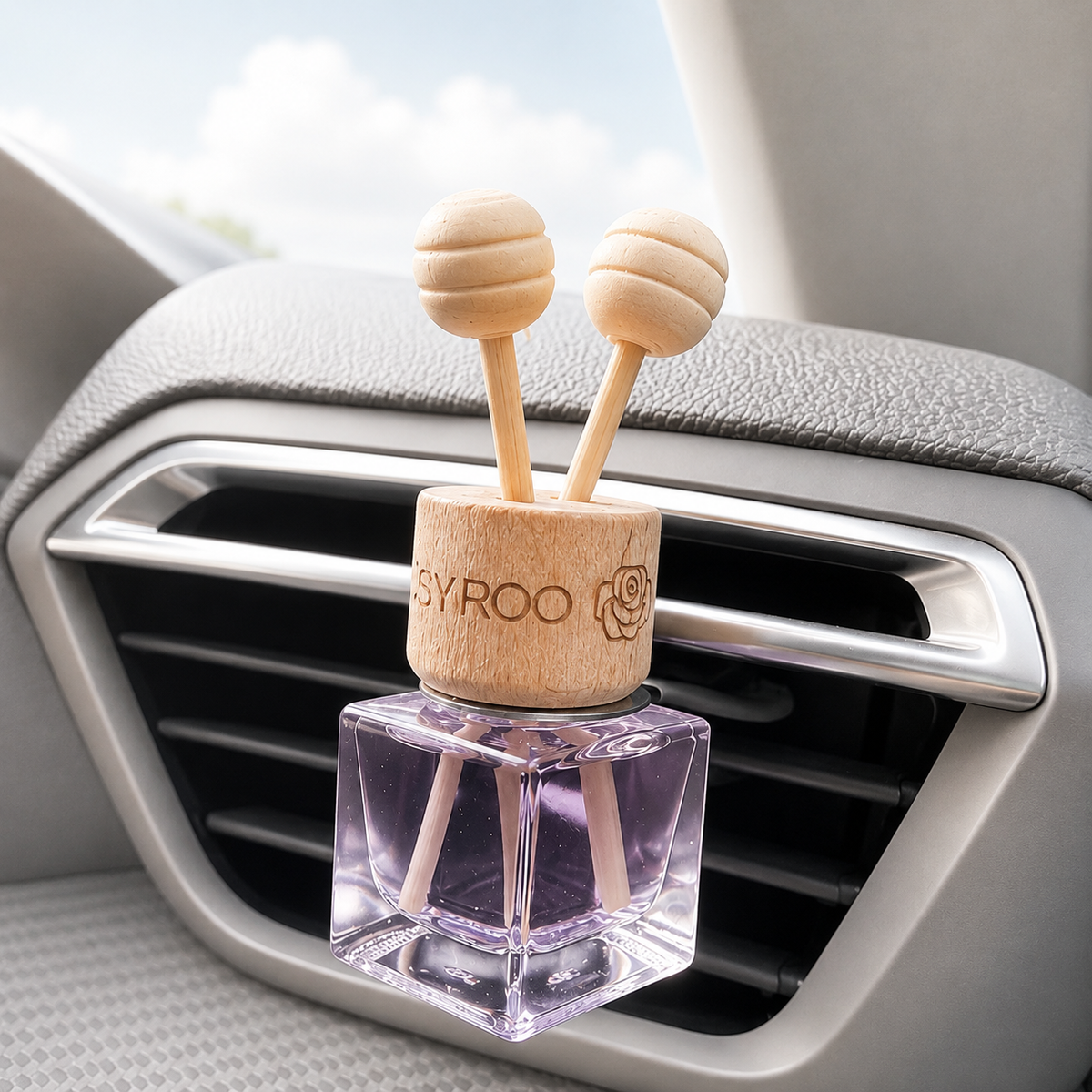 A car air freshener diffuser clipped onto a vehicle's air vent. The product features a clear square glass bottle filled with purple liquid, a light natural wood top with two wooden diffuser sticks, and a wooden collar engraved with the brand name 'SYROO'.