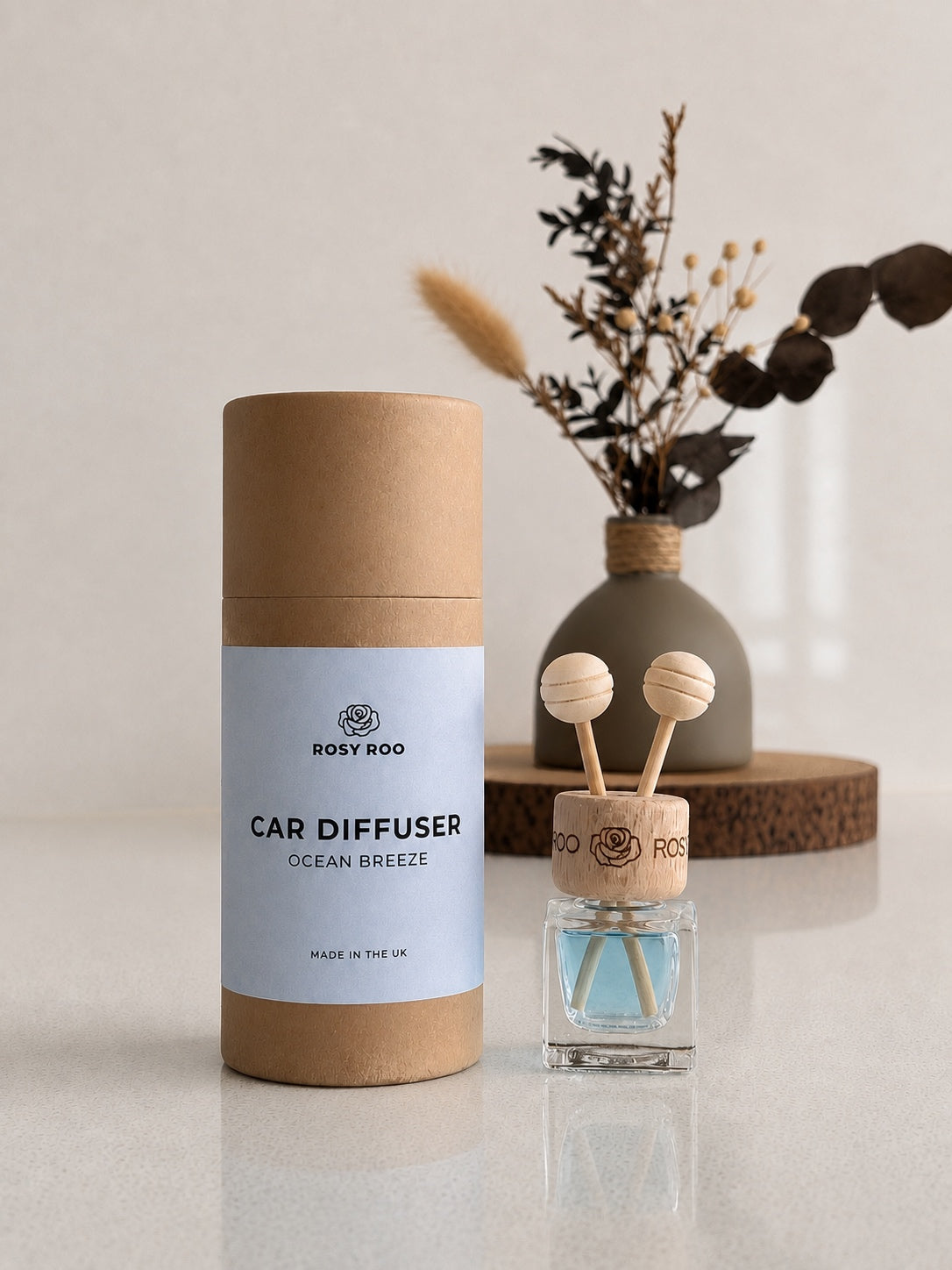 Rosy Roo Car Diffuser 'Ocean Breeze' shown with its brown cardboard packaging cylinder and the clear glass diffuser bottle with wooden diffuser balls and blue liquid, set on a light gray reflective surface with a neutral background and dried flowers.