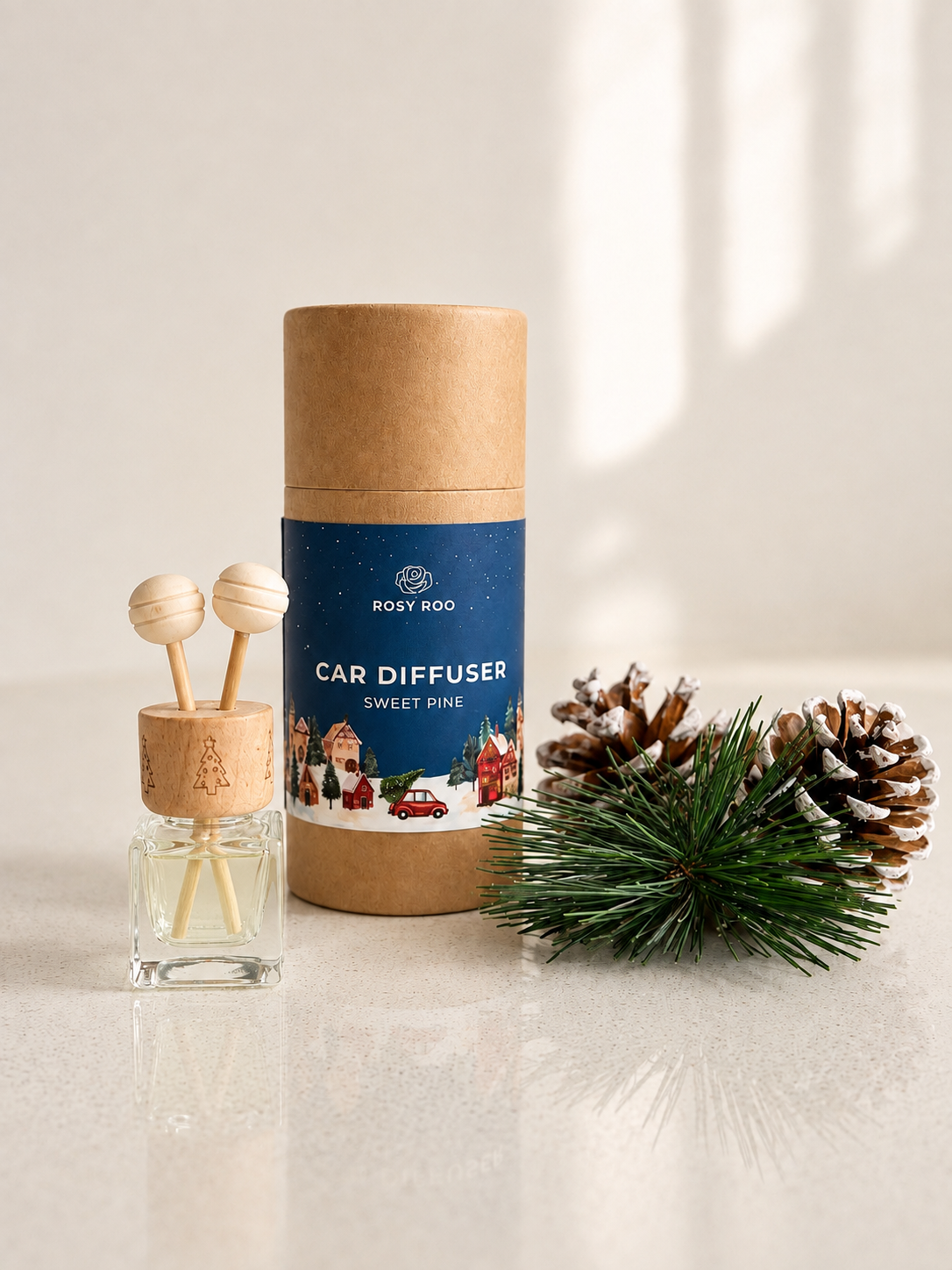 A car diffuser set by Rosy Roo in the Sweet Pine scent, featuring a kraft paper cylinder package with a blue label, a glass bottle with two wooden diffuser spoons, and pinecones and greenery on a light surface.